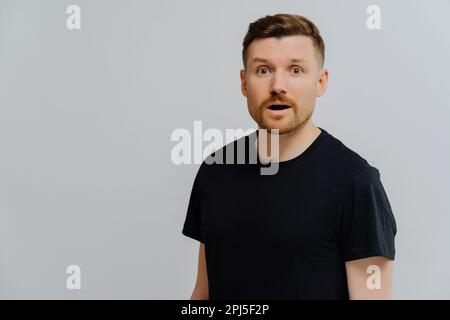 Studio shot of surprised intrigued handsome ginger man in black tshirt gazing with amazement at camera while standing shocked over gray background, co Stock Photo