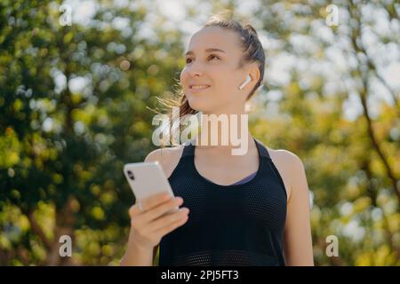 Thoughtful cheerful woman with pony tail has walk outdoor listens music while going in for sport holds modern smartphone leads healthy lifestyle focus Stock Photo