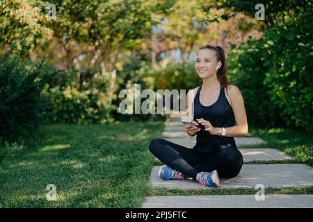 Cheerful female runner poses in lotus pose outdoor dressed in sportswear uses mobile phone during rest after jog downloads music songs in player prepa Stock Photo