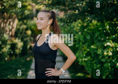 Half length shot of sporty young woman dressed in sportswear keeps hands on waist has dark hair combed in pony tail focused into distance with happy s Stock Photo
