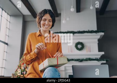 Christmas time. Happy young Spanish woman engaged in packing Xmas gifts, orders for customers, friends, enjoying winter holidays at home. Smiling euro Stock Photo