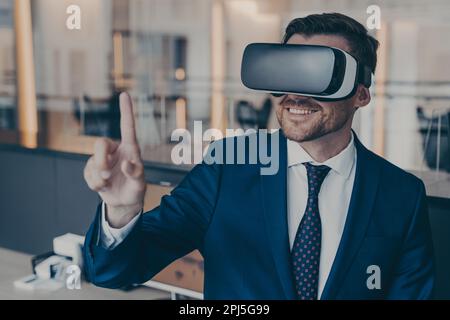 Close-up photo of smiling young bearded finacier in formal suit enjoying spending time in virtual reality with VR headset glasses, pointing with foref Stock Photo