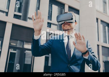 Young cheerful businessman with beard dressed in blue formal suit trying out VR glasses, checking out virtual reality, exploring digital world, standi Stock Photo