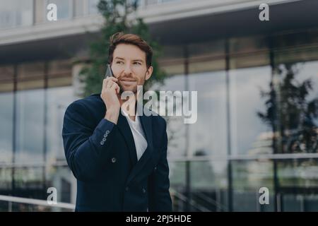 Male entrepreneur talks via phone solves urgent problems poses near business center dressed in black suit uses modern technologies for communication l Stock Photo