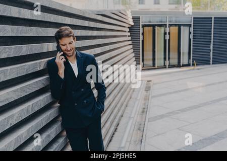 CEO male in smart casual having pleasing phone talk standing outside of office center entrance, leans on marbale striped facing outer wall, having cov Stock Photo