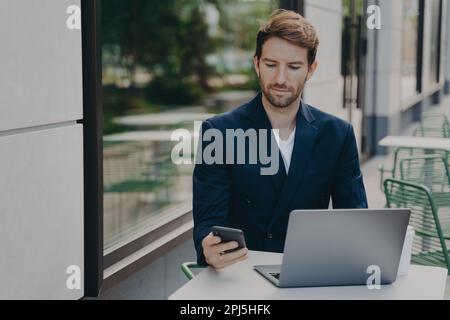 Serious male boss dressed in elegant clothes works online on laptop computer types text messages via smartphone consults clients checks email poses in Stock Photo