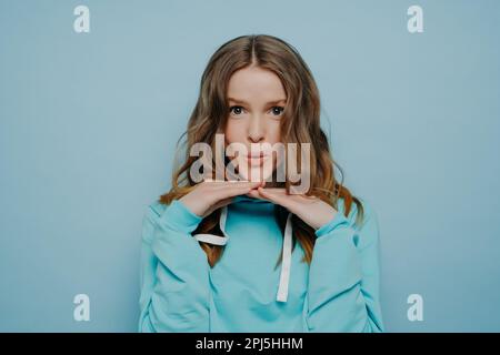 Pretty young teenage girl with wavy brown hair and widely open dark eyes touching her chin with fingertips expressing amusement standing on light blue Stock Photo