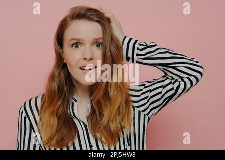 Attractive young female in stripy black and white blouse touching head with one hand in astonishment expressing surprise while posing against pink stu Stock Photo