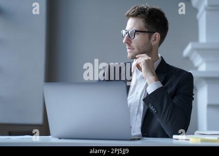 Focused young German male office worker sits at desk at home workplace distracted from laptop screen, pensive businessman looking aside immersed in th Stock Photo