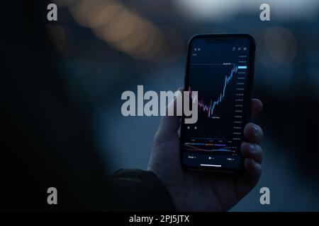 Broker or investor using stock trading app on smartphone to analyzing price flow on stock market while standing outdoors. Selective focus on hand hold Stock Photo