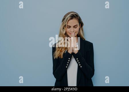 Beautiful young blond girl standing in praying pose folded palms together with her eyes closed, pleading and making wish come true, isolated over blue Stock Photo
