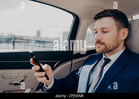 Male bearded investor in blue suit checking phone messages and notifications, receiving troubling news about his business, thinking and deciding his n Stock Photo