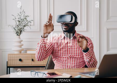 Focused Aframerican man in VR glasses enjoying augmented reality while sitting at workplace at home office, pointing with fingers up in air interactin Stock Photo