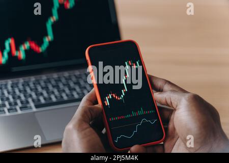 Male hands of African race holds smartphone in front of laptop, broker trader analyzing financial data graphs on phone and computer screen for investm Stock Photo