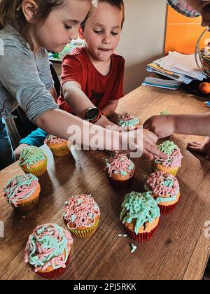 Group of children baking cupcakes, preparing ingredients, topping, sprinkles for decorating cookies. Kids cooking, working together in kitchen at home Stock Photo