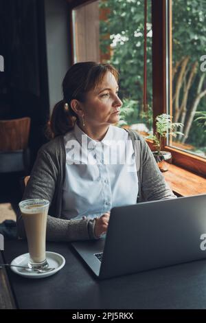 Focused businesswoman working remotely on her laptop computer contemplate managing her work sitting at the table in a cafe Stock Photo