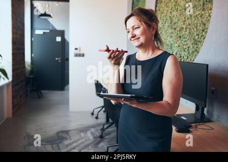 Businesswoman having business call working on tablet in office. Mature busy woman using touch pad computer standing by window in modern interior. Fema Stock Photo