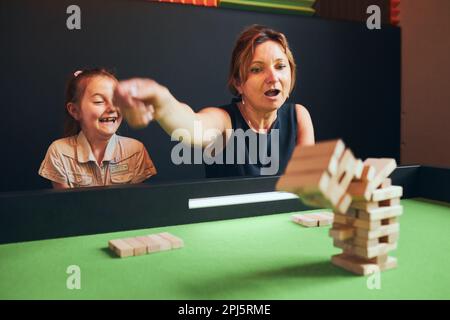 Excited woman playing jenga game with her daughter in play room. Woman removing block from stack. Falling tower of wooden blocks. Game of skill and fu Stock Photo