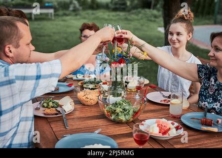 Family making toast during summer picnic outdoor dinner in a home garden. Close up of people holding wine glasses with red wine over table with food a Stock Photo