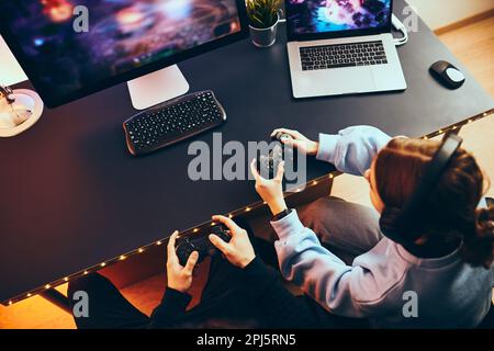 Friends playing video game at home. Gamers holding gamepads sitting at front of screen. Streamers girl and boy playing online in dark room lit by neon Stock Photo