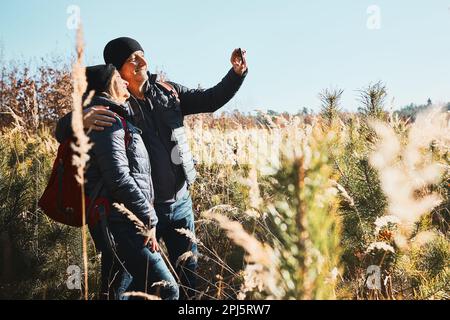 Hugging couple taking selfie while vacation trip. Hikers with backpacks on way to mountains. People walking through tall grass along path in meadow on Stock Photo