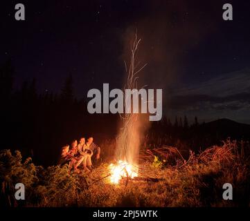 Happy family sitting near camp fire and warming up. Cozy atmosphere at starry night in mountains. Big family spending time together on hike. Stock Photo