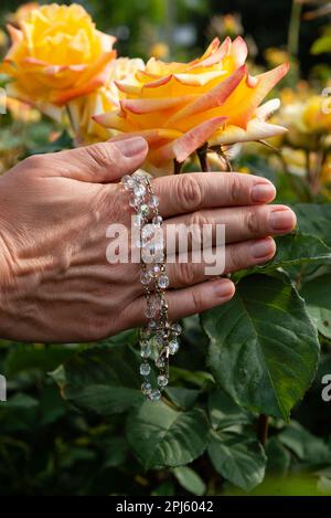 Cropped view of female hands holding rosary with glass beads on yellow roses background, selective focus. Stock Photo