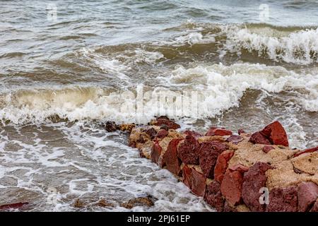 Sea water breaking over a small uneven reddish-brown stone wall on the beach, forming abundant foam, rocky coast, irregular surface, cloudy day Stock Photo