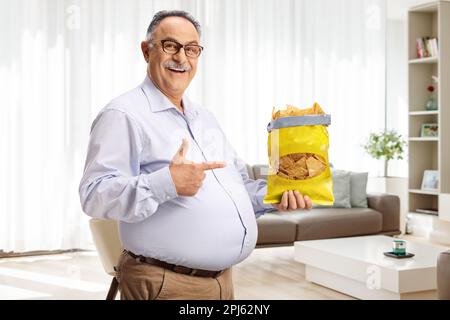 Cheerful mature man holding a pack of tortilla chips and pointing at home in a living room Stock Photo