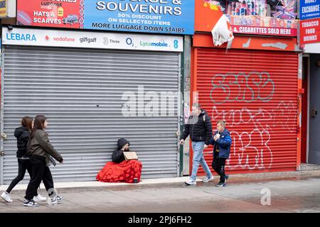 Woman begging sits on the pavement under a sleeping bag outside a closed down shop with its shutters down while people pass by on Oxford Street on 26th March 2023 in London, United Kingdom. The scene is illustrative of the social disparity in the UK with some people who live in relative wealth in comparison to others. Stock Photo