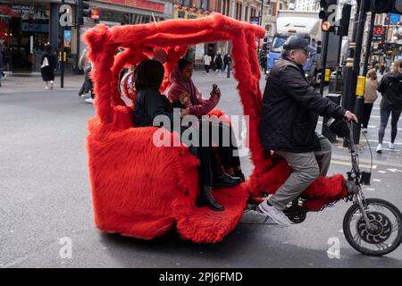 Two passengers take a cycle taxi ride in a red fake fur rickshaw in Soho on 27th March 2023 in London, United Kingdom. Stock Photo