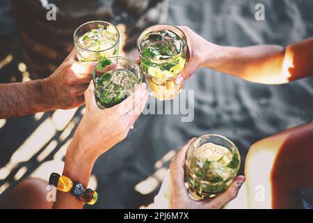Four friends toasting with mojito glasses while sitting at a beach bar in the summer - hands holding drinks on sandy beach. Stock Photo