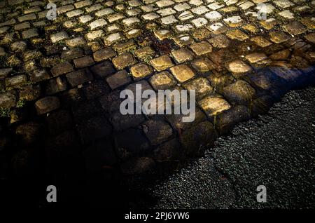 Old cobble stones and tarmac road surface under a puddle. Stock Photo