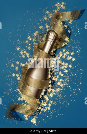 Luxury composition with golden champagne bottle and decorations of golden star shaped confetti and party streamer on blue background in flat lay style. Christmas or New Year Eve celebration concept. Stock Photo