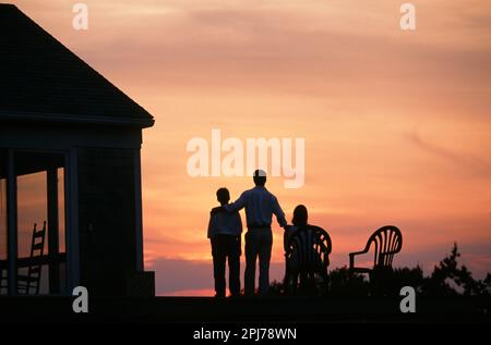 Family of three silhouette it against a beautiful sunset oh relaxing on the deck of their Home Stock Photo