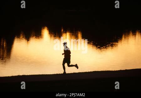 A silhouette of a man jogging alone a leak at sunset Stock Photo