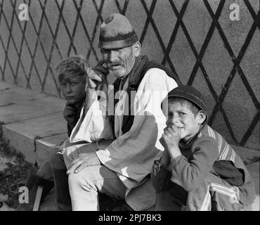 Sibiu County, Socialist Republic of Romania, approx. 1977. Shepherd wearing traditional clothing and children on a sidewalk in the city. Stock Photo