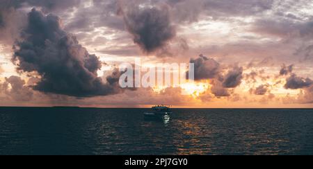 A stunning sunset low-key panoramic view of a boat in the sea. The orange sun is partially hidden behind violet and pink clouds; The boat's silhouette Stock Photo
