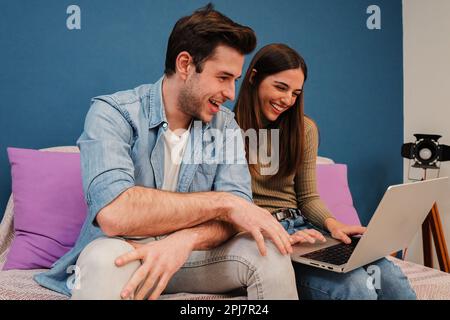 Caucasian young couple browsing on internet using a laptop compuer sitting on a sofa in their home. Happy friends smiling and having fun watching videos on a social media at their apartment indoors. High quality photo Stock Photo