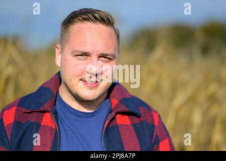 Young man with a red and blue lumberjack jacket and blue t-shirt in front of an autumnal corn field as a background Stock Photo