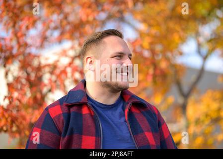 Young man with a bright smile in a peasant jacket stands in front of an autumn background with trees and yellow leaves Stock Photo