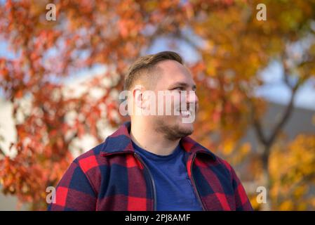 Young man with a bright smile in a peasant jacket stands in front of an autumn background with trees and yellow leaves Stock Photo