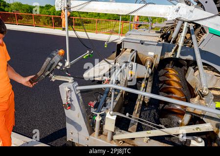 Operator, worker is holding industrial remote control, console with joystick to directing asphalt spreading machine, determines position for pouring, Stock Photo