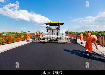 View on machine for laying asphalt, spreading layer of hot tarmac on prepared ground a few workers are using shovels to level it. Stock Photo