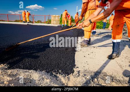 Few workers are using shovels to level, set up layer of fresh tarmac to right measures, pouring hot asphalt. Stock Photo