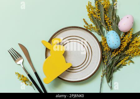 Easter table setting. Plate with easter eggs and mimosa on a pastel green background. Happy Easter holiday concept for restaurants. View from above. Stock Photo