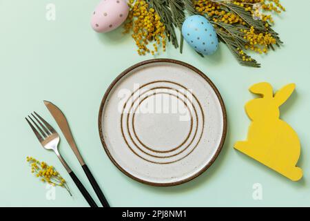 Easter table setting. Plate with easter eggs and mimosa on a pastel green background. Happy Easter holiday concept for restaurants. View from above. Stock Photo