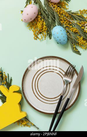 Easter table setting. Plate with easter eggs and mimosa on a pastel green background. Happy Easter holiday concept for restaurants. View from above. C Stock Photo
