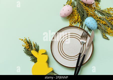 Easter table setting. Plate with easter eggs and mimosa on a pastel green background. Happy Easter holiday concept for restaurants. View from above. C Stock Photo