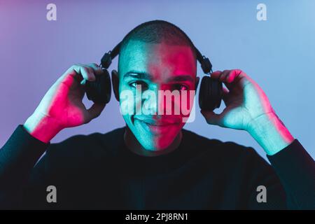 Music in life concept. Positive young adult man looking to the camera with a smile and putting on his black wireless headphones. Closeup portrait. High quality photo Stock Photo